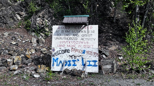 A handwritten sign on a gravel road reads “Road Closed” and welcomes family and friends to Madii Lii, with rocky terrain and trees in the background.