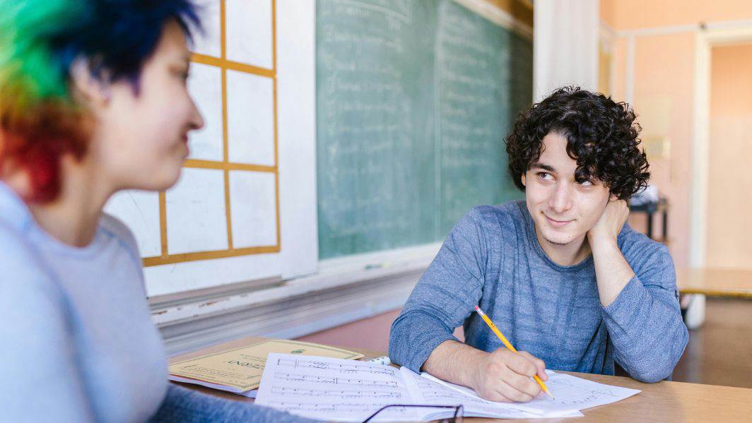 A student sits at a desk, focused on writing in a notebook, with notes and a pencil in hand, surrounded by a classroom setting.