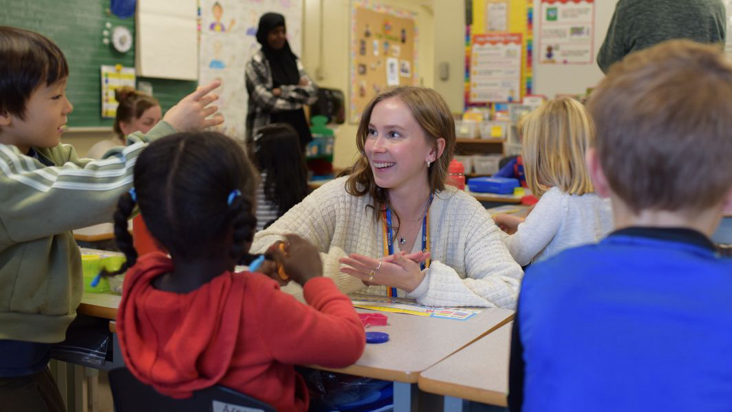 A young teacher is pictured in an elementary classroom, crouched in front of a desk at eye-level with a kindergarten student. She is looking at the student and offering encouragement.