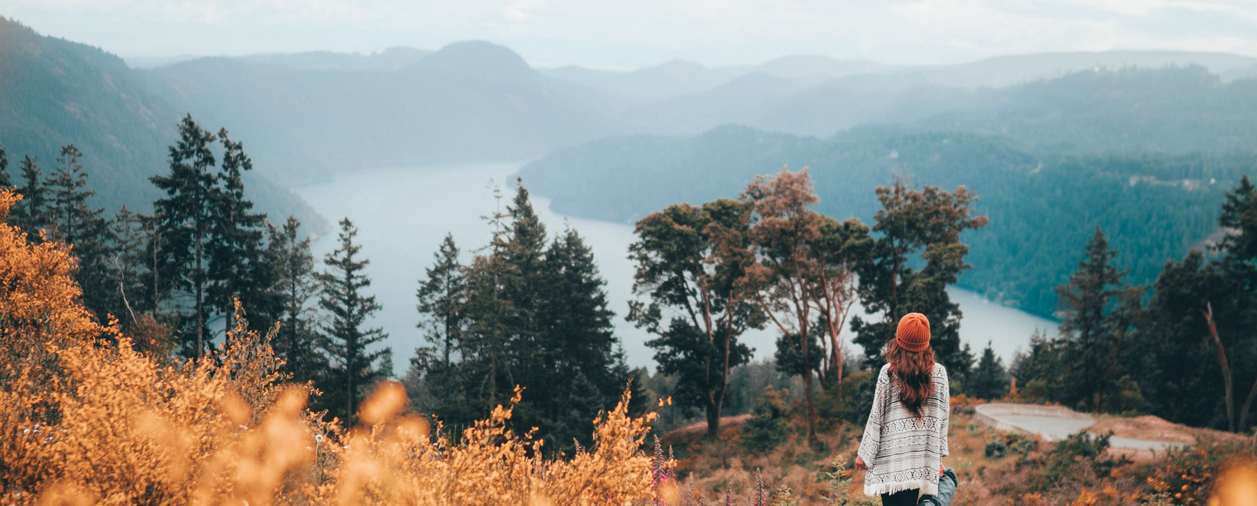 A hiker enjoying the view of a tranquil ocean inlet from a hilltop, with surrounding mountains.