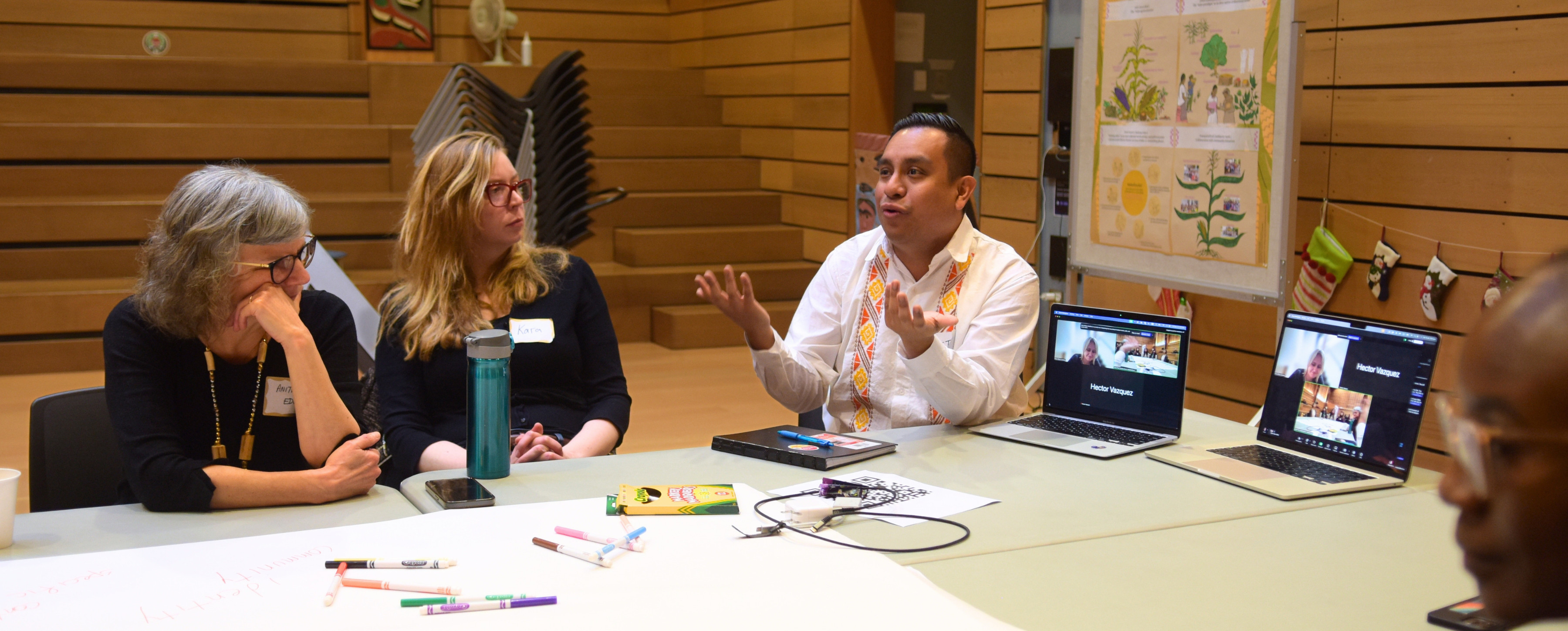 Four people engaged in a discussion around a table with laptops and papers. A man gestures expressively while others listen attentively.