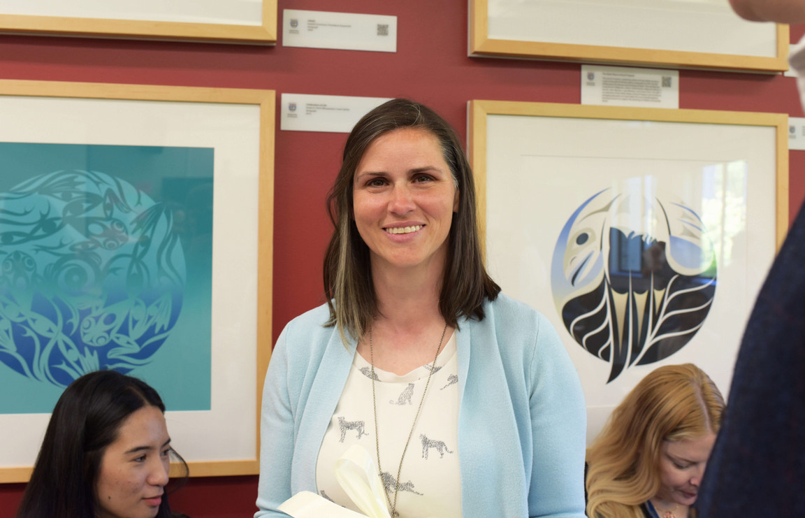 Smiling woman in light blue cardigan holds a notebook in an art gallery. Colorful abstract artworks with nature motifs hang on a red wall. Warm, welcoming atmosphere.