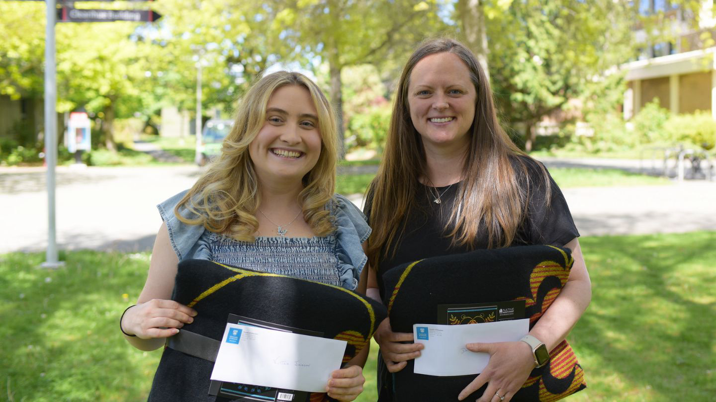 Two Faculty of Education 2024 award winners (Victoria Jackson and Jean Buckler) pictured together outside the MacLaurin Building, each holding a folded blanket and award envelope.