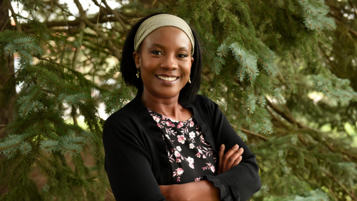 A portrait of Dr. Doris Kakuru. She is pictured outdoors, wearing a black blazer, black floral patterned top, and light brown headband. She is smiling at the camera with her arms crossed.
