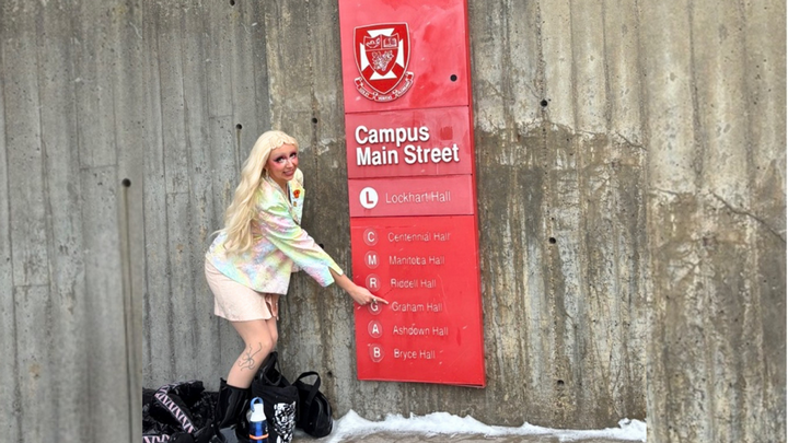 EKTEP student Graham Smith stands in front of a university sign reading 'Graham Hall'. They are in drag and preparing to present at the WestCAST conference. They are wearing a white and pink sparkly skirt suit and long blonde hair.
