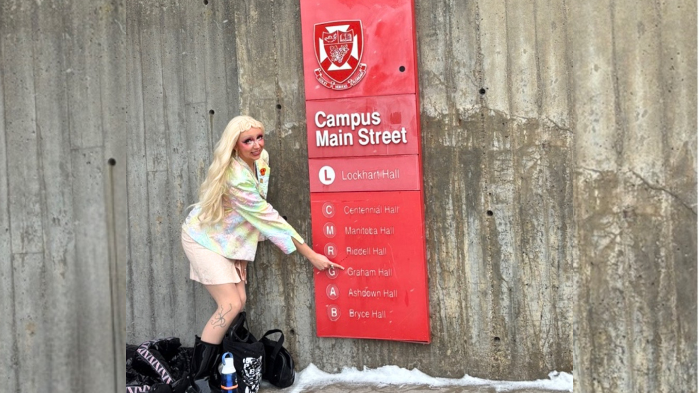 EKTEP student Graham Smith stands in front of a university sign reading 'Graham Hall'. They are in drag and preparing to present at the WestCAST conference. They are wearing a white and pink sparkly skirt suit and long blonde hair.
