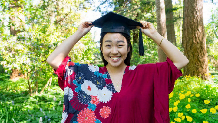 An international student is pictured in Finnerty Gardens, smiling, wearing a dark red dress and their graduation cap.