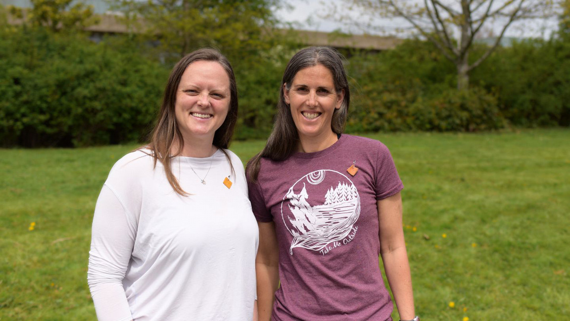 Two women smiling outdoors on a sunny day. One wears a white shirt, the other a purple shirt with a nature design. Background shows green grass and trees.