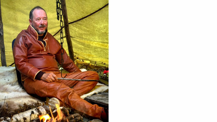 A man in a cozy brown outfit sits cross-legged on a fur rug inside a tent, smiling warmly. A small fire burns in front of him, creating a rustic, peaceful ambiance.