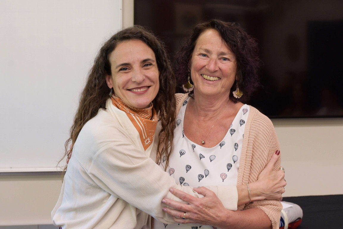 Two women smiling warmly and embracing in a classroom setting. The woman on the left has long curly hair and wears a cream-colored top with a rust-colored scarf, while the woman on the right has short dark curly hair and wears a pink cardigan over a white shirt with balloon prints.