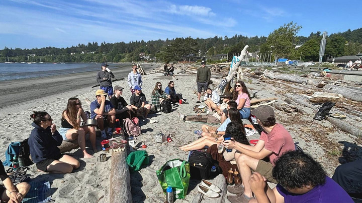 A group of students chilling together, sitting on blankets on the beach on a bluebird day. 