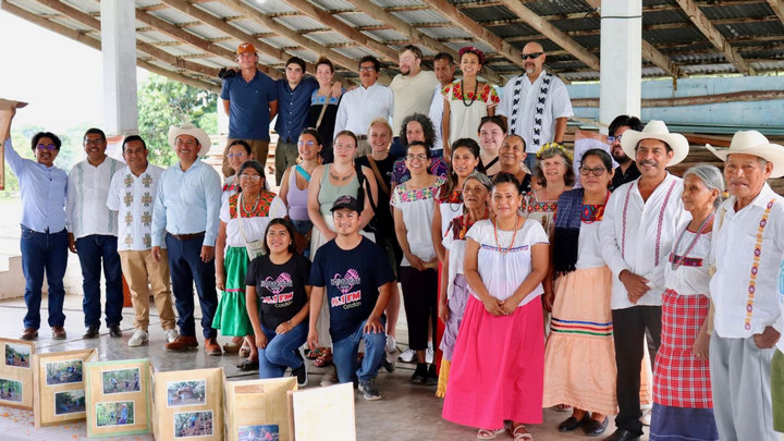 A diverse group of people pose together indoors, wearing colorful, traditional clothing and casual attire. Wooden boxes with photos are in the foreground. The mood is joyful and celebratory.