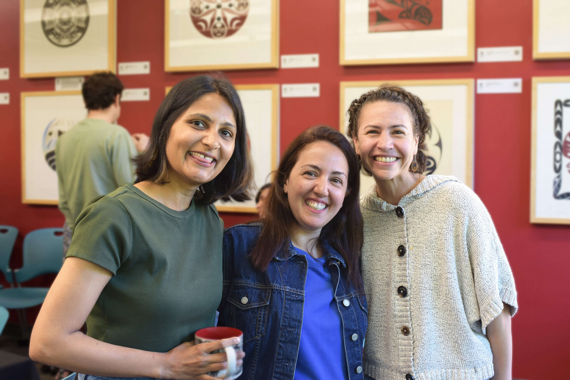 Three women smiling warmly in a gallery with framed artwork on red walls. One holds a mug, suggesting a relaxed, cheerful gathering.