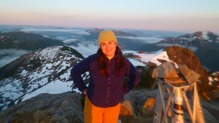 Olivia is pictured in a blue jacket and yellow beanie stands atop a snowy mountain at sunrise, smiling with hands on hips, surrounded by a vast landscape of peaks and clouds.