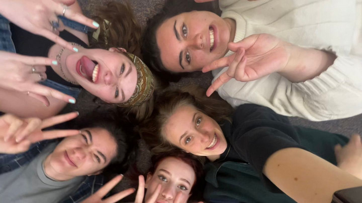 Five friends lying on the floor in a circle, joyfully smiling and making peace signs. The atmosphere is fun and energetic, with a sense of camaraderie.