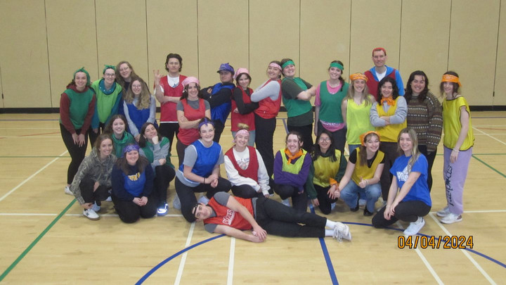 A group of smiling people in colourful sports jerseys and hats pose happily in a gym. The floor has blue and white lines. Dated April 4, 2024.