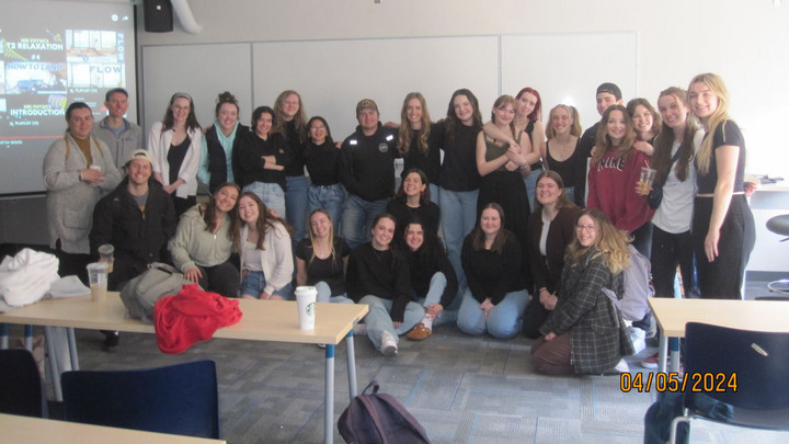 A group of smiling young adults pose in a classroom, suggesting a celebratory or end-of-semester vibe. The mood is cheerful and relaxed.