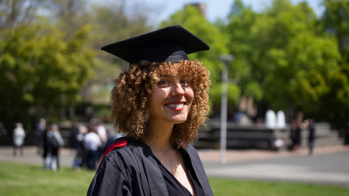 A student is pictured on campus in their graduation cap and gown. They are smiling toward the camera.