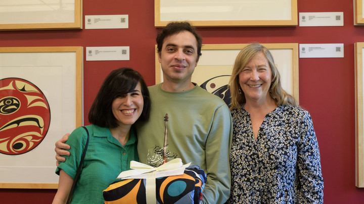 Three people smiling in front of framed Indigenous artwork on a burgundy wall. One person holds a gift wrapped in a colorful, patterned fabric.