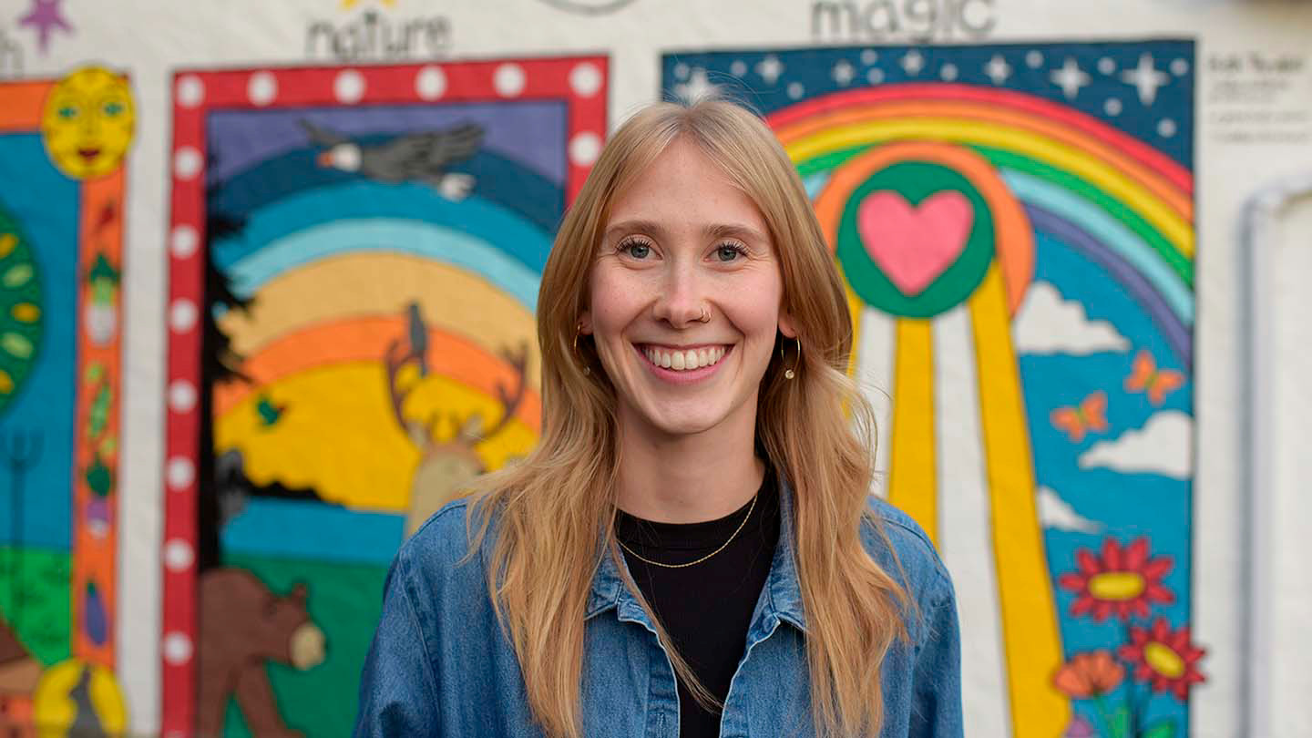 A smiling young woman with long blonde hair wearing a denim jacket stands in front of a colorful mural. The mural features vibrant imagery including a rainbow, a heart, a deer, and flowers, with the words "nature" and "magic" partially visible at the top.