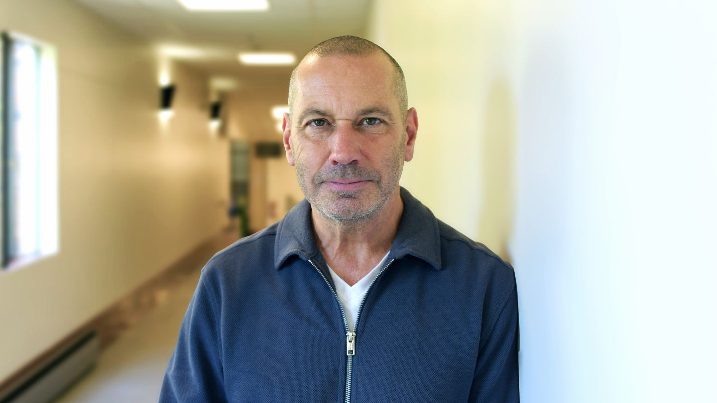 A portrait of Dr. Todd Milford. He is pictured in a hallway in MacLaurin Building, wearing a navy blue knitted sweater and looking into the camera lens.
