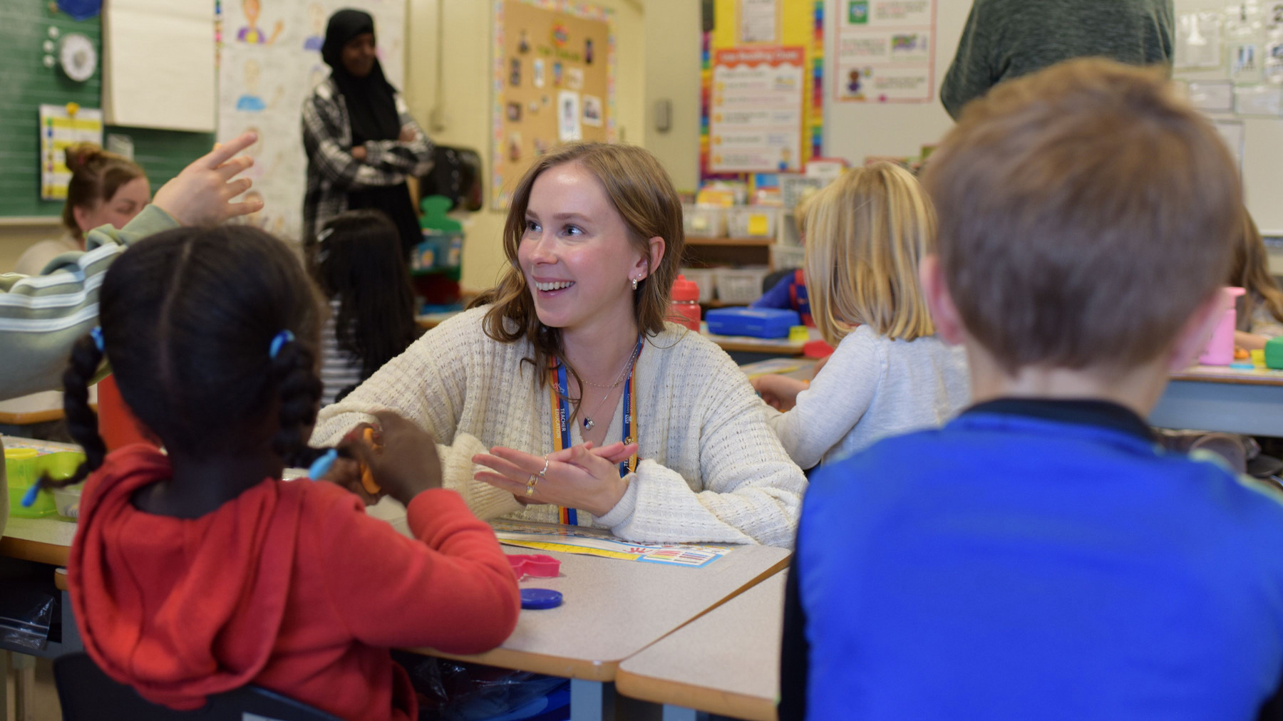 A teacher with a warm smile interacts with a group of young children in a brightly lit classroom. Children sit around a table engaged in activities, with one girl in a red hoodie at the forefront and another child raising their hand.