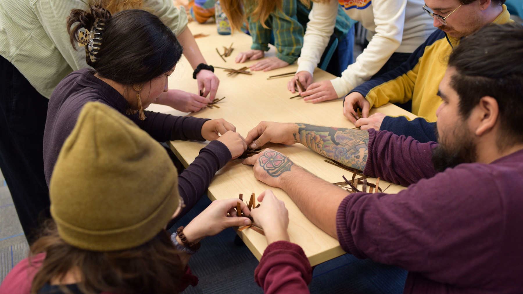 A group of students gathered around a table, taking part in a cedar weaving activity with visiting instructor Tyrone Elliott.