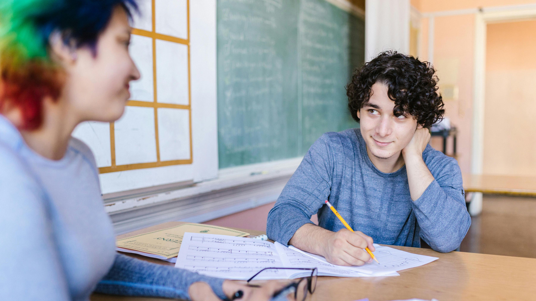 Two students are seated across from another at a table, talking and taking eye contact with each other. One student is facing the camera and holding a pencil, the other is facing away and has colourful hair.