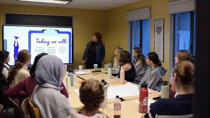 A group of CORE tutors seated around a table listening to a presentation. The presenter has a slideshow and is training the tutors on working with neurodiverse students and colleagues.