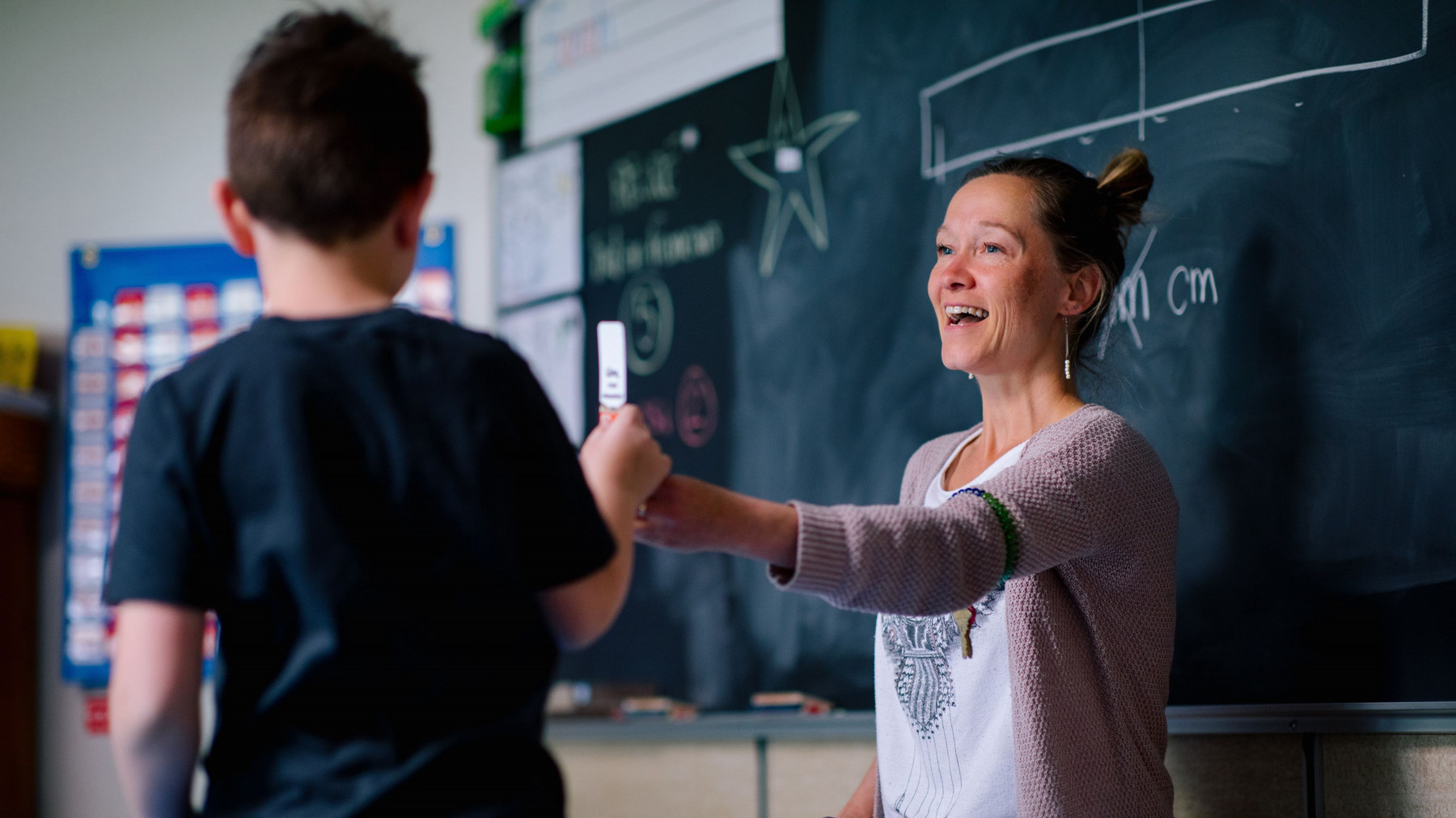 A teacher with a warm smile interacts with a student who is facing away from the camera. The teacher is handing the student a piece of chalk with an encouraging mood.