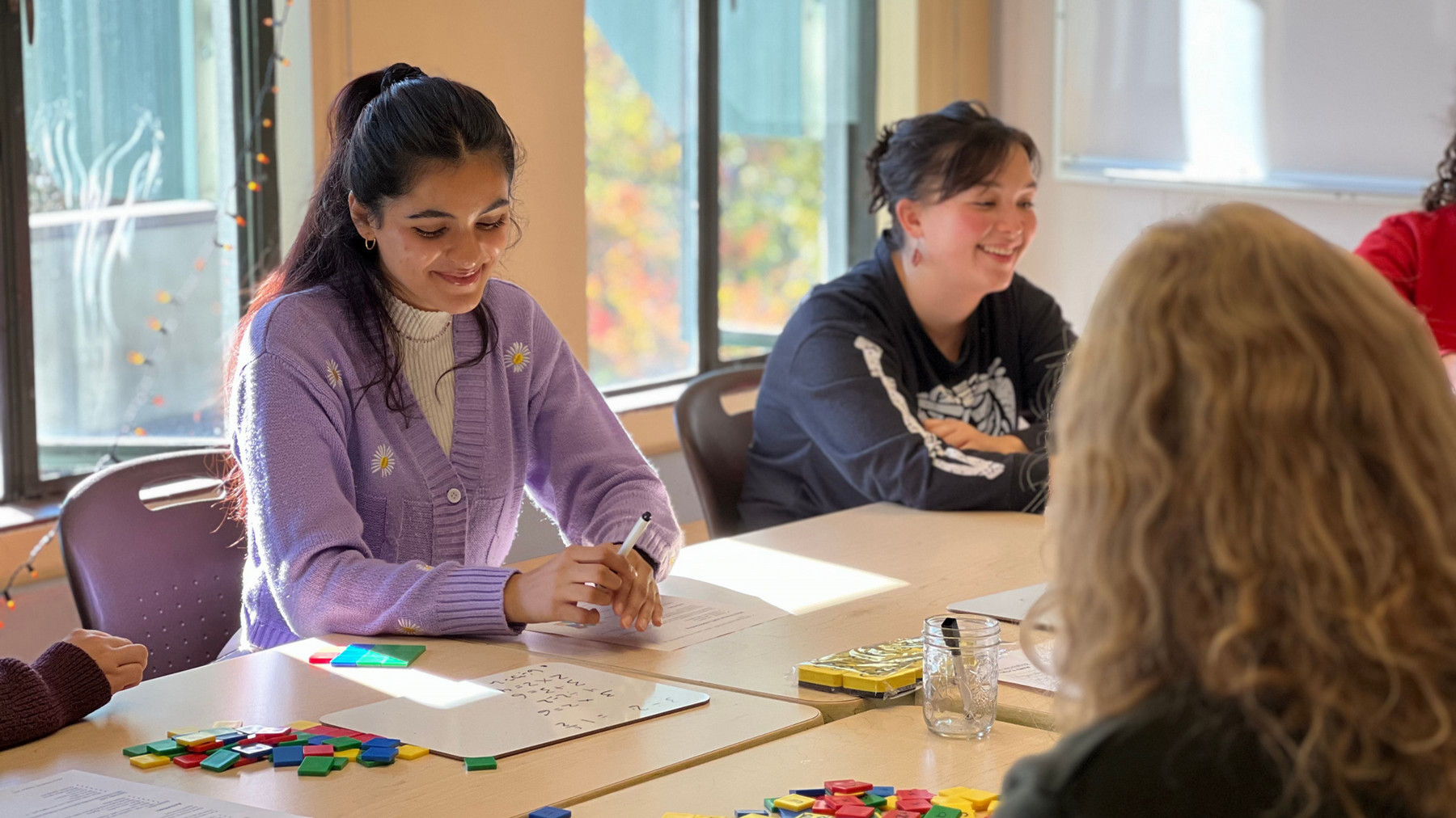 A student sits at a table in the CORE learning centre on a sunny day, with light streaming in through a window behind them. The student is smiling and working on math activities on the tabletop.