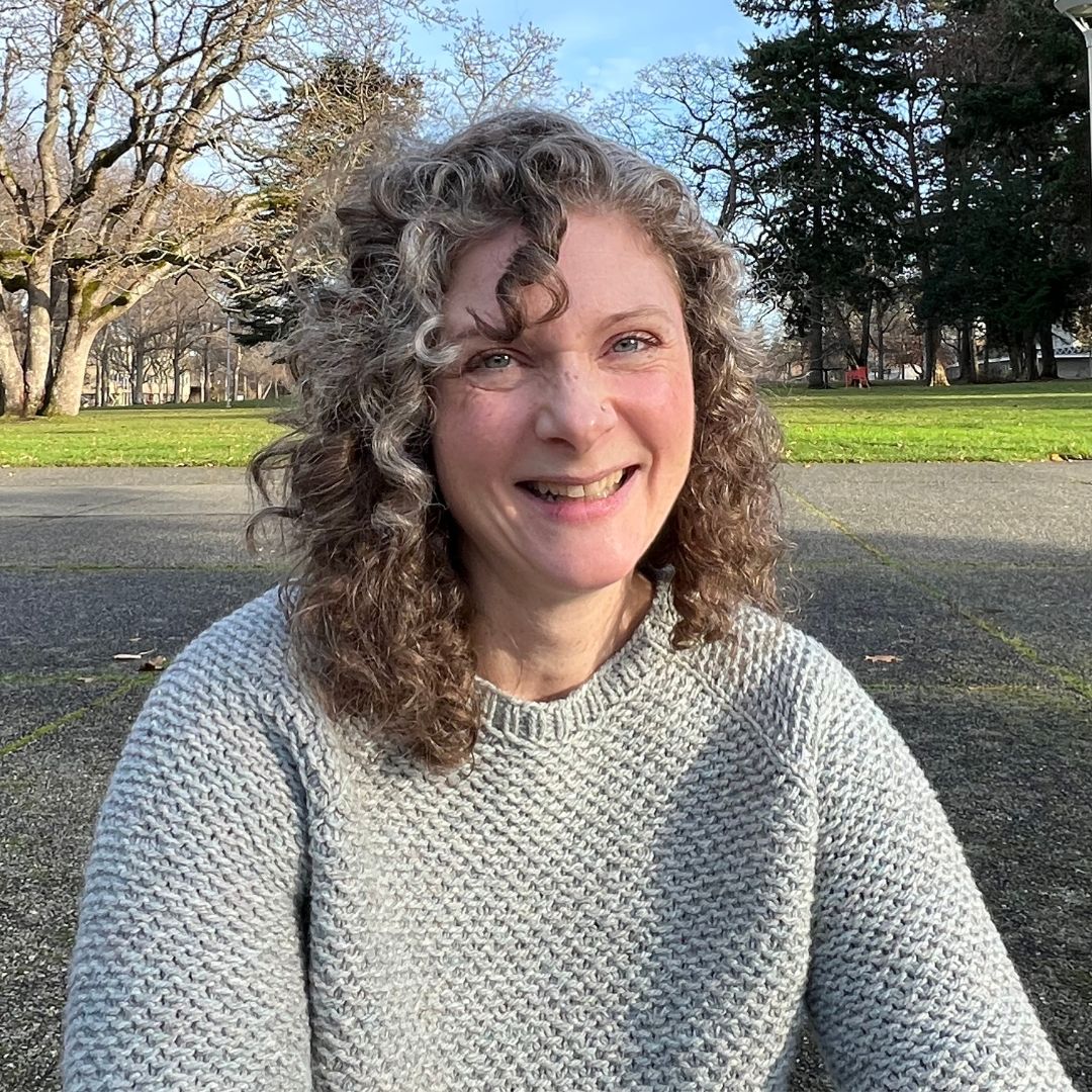 A portrait of Gillian outdoors on campus. She is smiling at the camera, wearing a grey cable knit sweater.