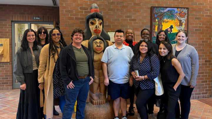 11 PhD students standing in front of the Transformation Pole outside the School of Child and Youth Care office.