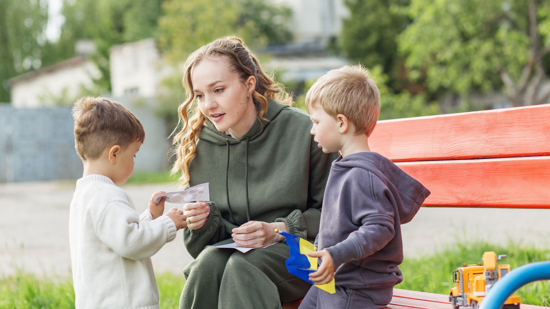 A woman in a green hoodie sits on a red bench, engaging with two young boys. One boy holds toy trucks while the woman examines a card. The scene is outdoors, surrounded by greenery.