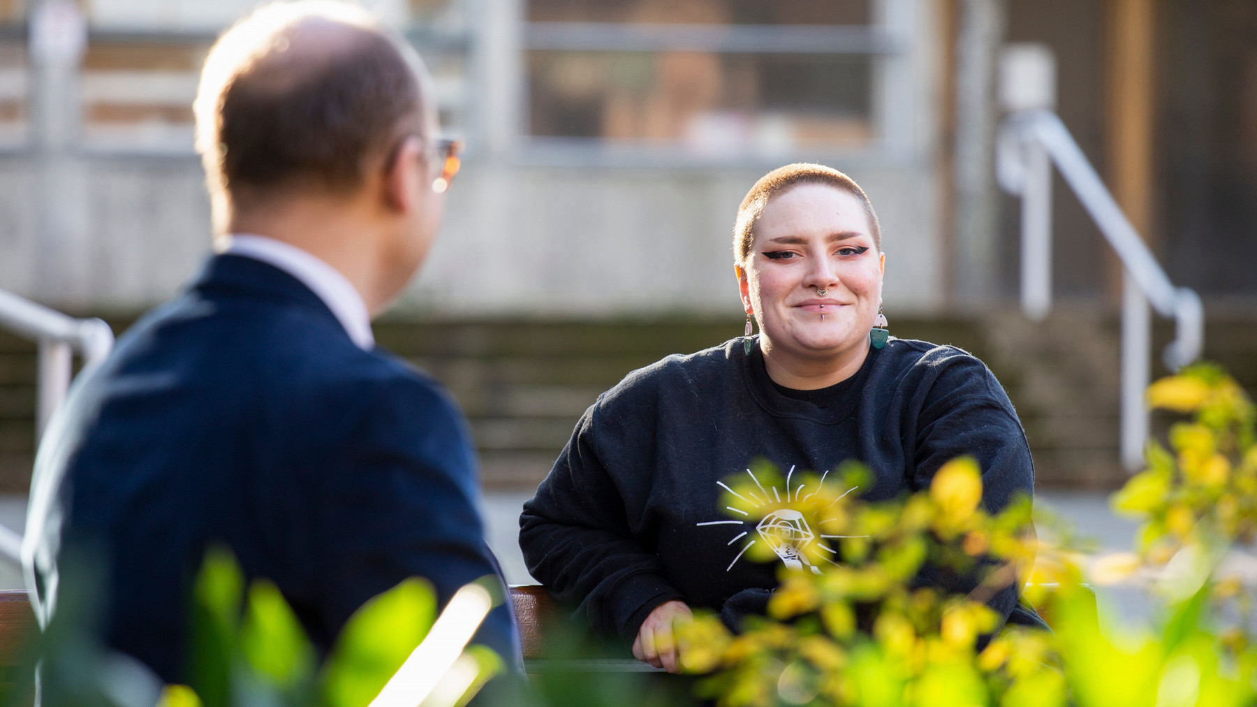 A person with a shaved head and earrings smiles warmly and confidently toward the camera, sitting outdoors. Another person, blurred in the foreground, listens attentively. Sunny, relaxed setting.