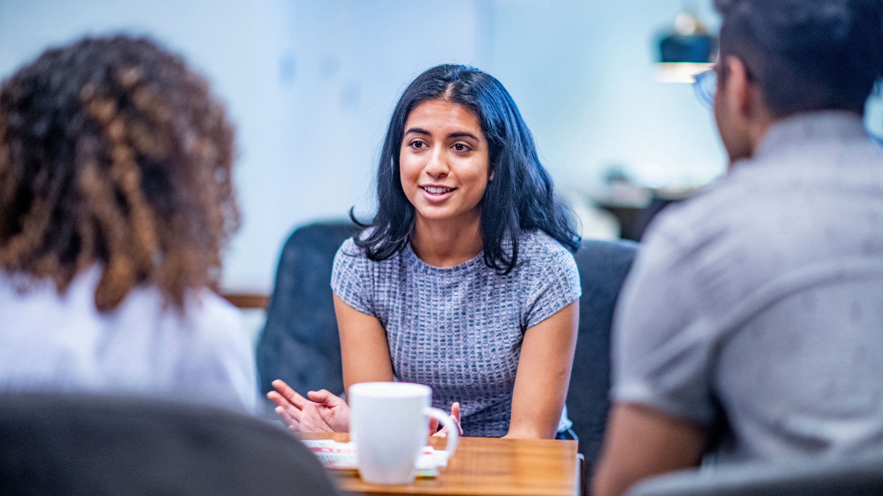A woman in a gray shirt engages in a conversation with two others in an office setting. A white mug is on the table, creating a collaborative, friendly tone.