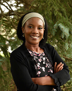 Doris Kakauru stands in front of an evergreen tree, smiling, arms crossed.