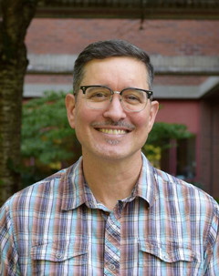A head-and-shoulders portrait of Jeff Smith on campus. They are pictured outdoors, wearing a plaid collared button-down shirt.