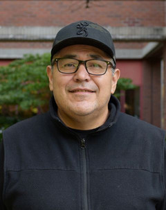 A head-and-shoulders portrait of Nick Claxton on campus. They are pictured outdoors, wearing a black sweater, glasses, and a black baseball cap.