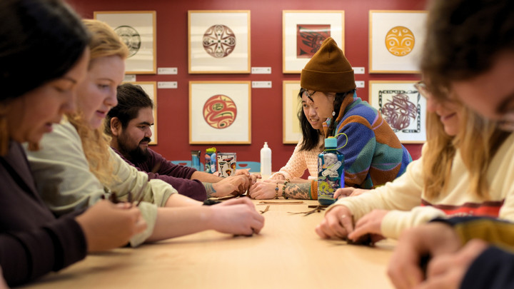 Students sit around a table, engaging in a cedar weaving activity. The room is adorned with Indigenous artwork, creating a focused and collaborative atmosphere.