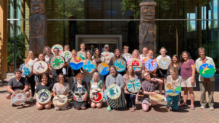 The group of summer institute students and instructors are pictured outside together on a sunny day in front of First Peoples House, many are holding painted drums (2022)