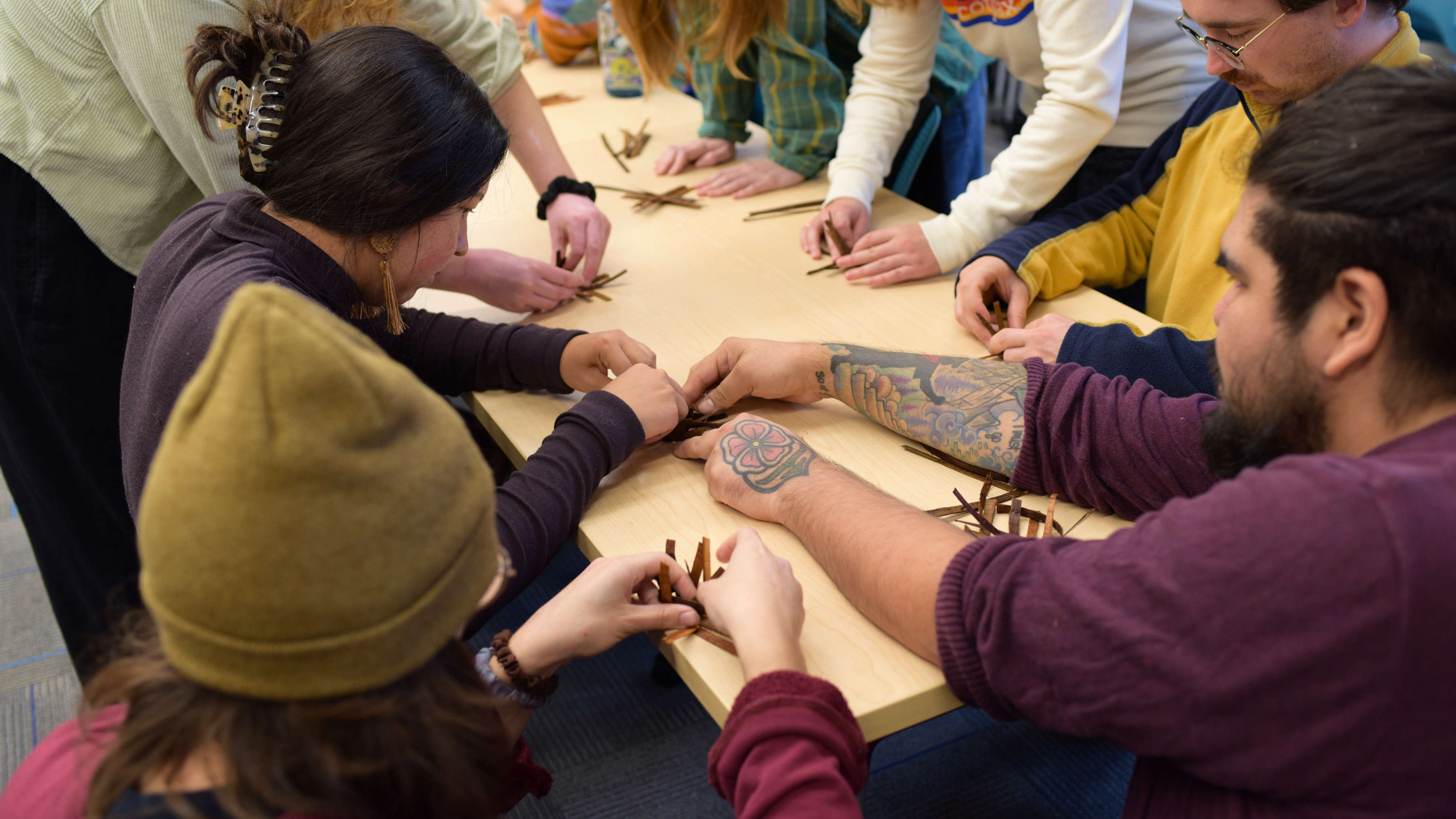 A group of people gather around a table engaged in a collaborative cedar weaving activity. The atmosphere is focused and cooperative.