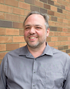 A portrait of Jean-Paul Restoule. He is pictured outdoors in front of a brick building. He is smiling, wearing a silver/grey collared button-down shirt.
