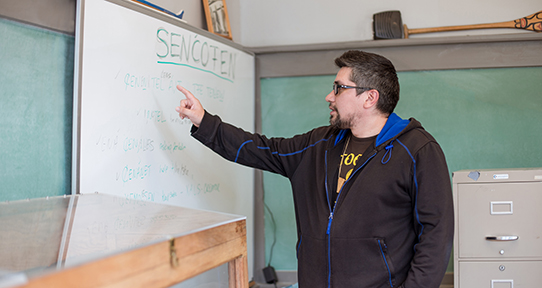 Man in a classroom pointing to a board with SENĆOŦEN words