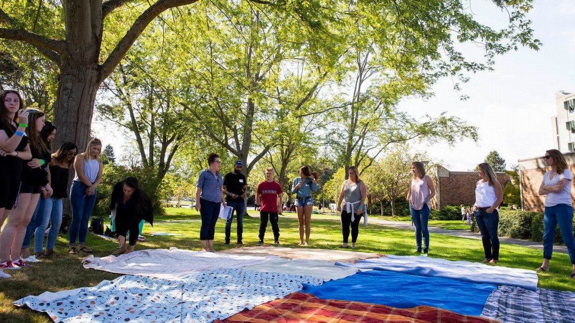 A group of students and an instructor standing outdoors, in circle, on campus, doing a blanket exercise. It's a bright sunny day.
