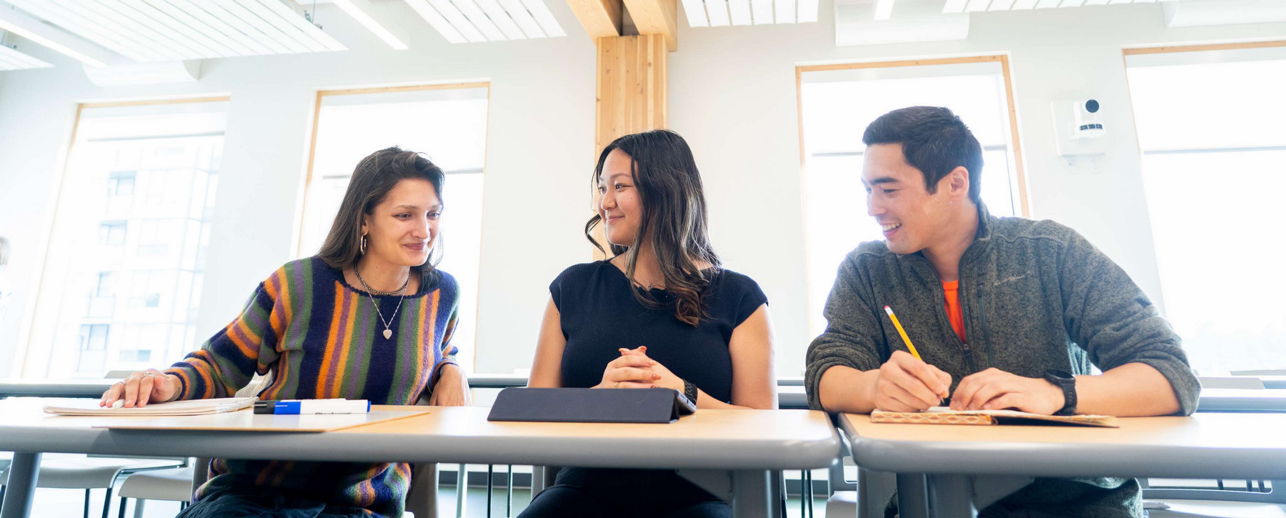 Three students sit together at a table in a bright, airy classroom, working together and chatting. On the table are two notebooks and an ipad.