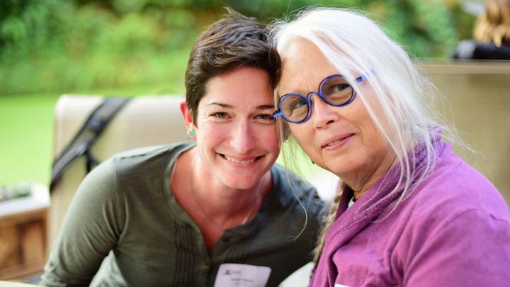 Two people sitting closely together and smiling at the camera outdoors, one with short dark hair and the other with long gray hair and blue glasses.