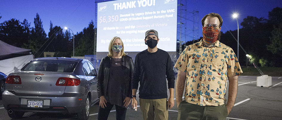 A family of three wearing masks, stand in a parking lot, in front of a parked car with a large movie screen in the background
