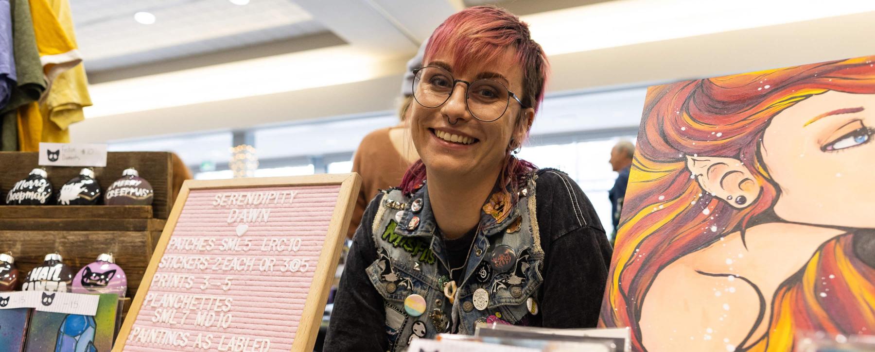 A woman with coloured hair and glasses smiles flanked by her artwork at the United Way Artisan's Market.