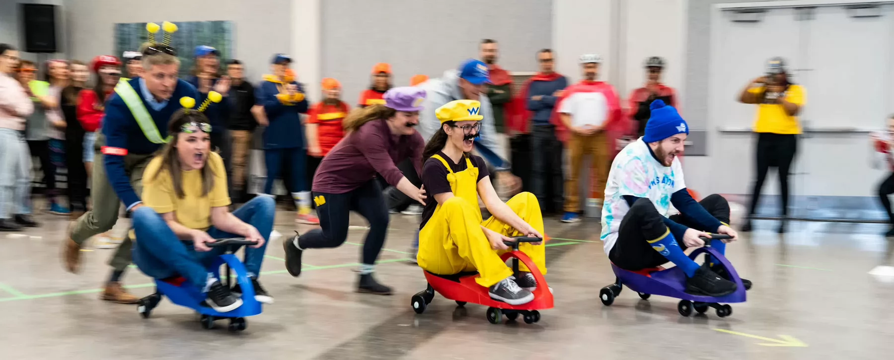 An action shot of a crowd of people cheering on teams of plasma car racers, each wearing costumes.
